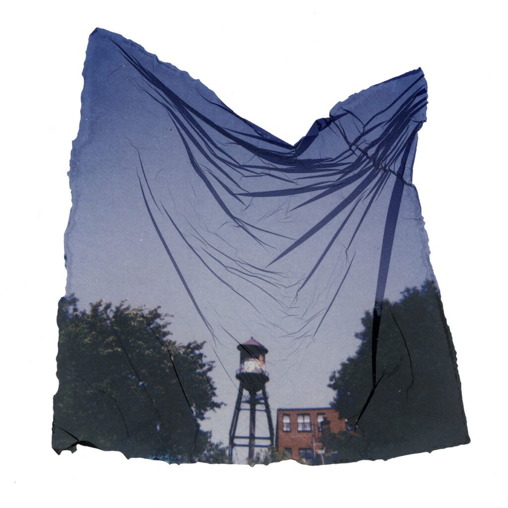 A water tower framed by two big trees sits in the bottom thiurd of the photo. The clear blue sky above shows many creases from the emulsion lift process. 